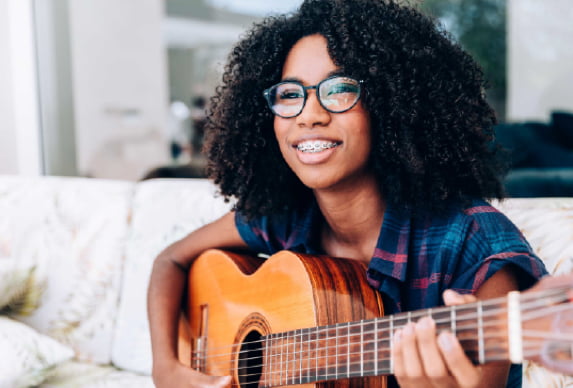 Person wearing glasses and holding an acoustic guitar while seated indoors.