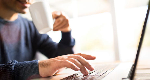 Person working on a laptop while holding a coffee mug at a desk