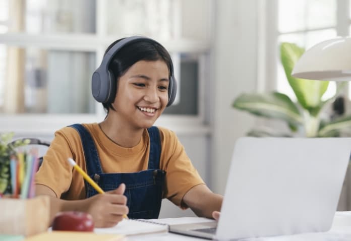 A smiling student wearing headphones and a denim jumpsuit sits at a desk, writing with a pencil in a notebook while looking at an open laptop, symbolizing flexible, self-paced online learning.