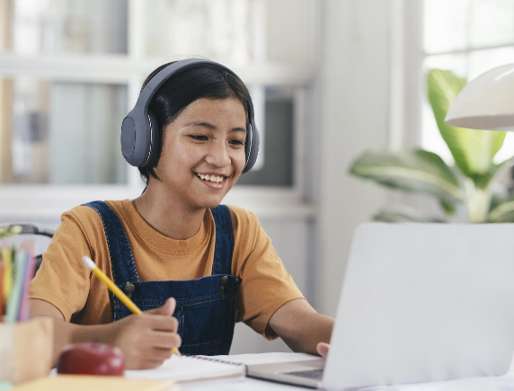 A smiling student wearing headphones and a denim jumpsuit sits at a desk, writing with a pencil in a notebook while looking at an open laptop, symbolizing flexible, self-paced online learning.