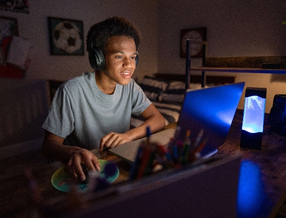 A focused high school student wearing headphones is sitting at a desk and looking at his laptop screen, representing a student engaged in online virtual school learning.