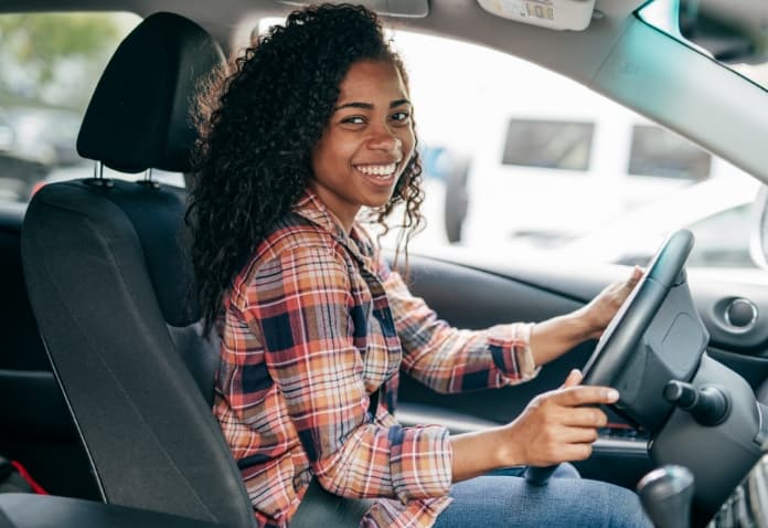 A smiling female student with curly hair is sitting in the driver's seat of a car with her hands on the steering wheel, ready to drive.