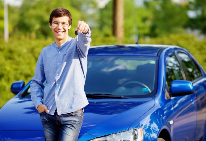 A grinning teenage boy is standing next to a blue car, holding up a set of car keys triumphantly.