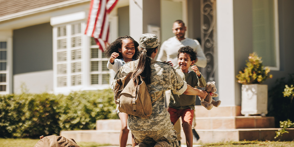 Military service member kneeling on a front lawn, embracing two children running toward them, with an American flag on the house.