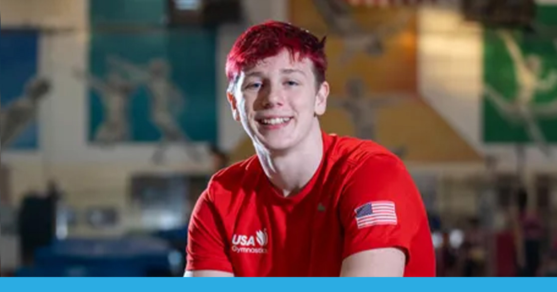 West Fowler wearing a red USA Gymnastics shirt with an American flag patch, seated in an indoor gymnastics facility with colorful wall murals in the background.