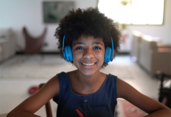 Student seated at a table wearing blue headphones and looking at a computer during a virtual learning session.