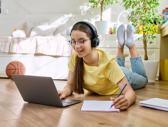 Female high school student enjoying the flexibility of online learning, working on her laptop and taking notes while relaxing on her bedroom floor.