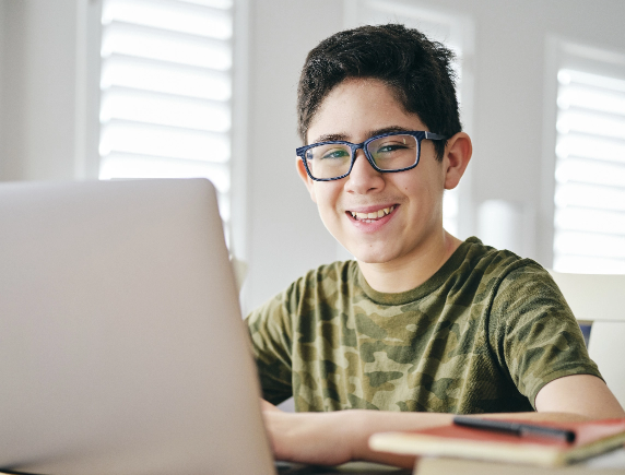 A smiling middle school boy wearing glasses works on his FLVS virtual school course from home using a laptop.