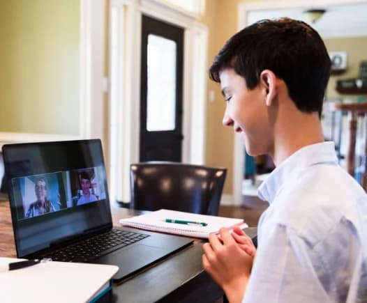 A male student in a light blue shirt smiles while participating in a virtual class video conference on his laptop, showing real-time interaction with his teacher and peers.