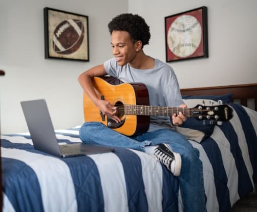 A smiling student sits on his bed, playing an acoustic guitar while viewing a laptop. This image symbolizes the flexibility of online school, allowing time for music and hobbies.