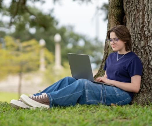 A female student with glasses and a dark shirt sits comfortably in the shade of a large tree trunk, working on her silver laptop in a grassy, outdoor setting.